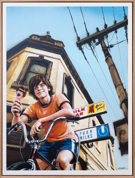 A framed art print depicting a young boy in an orange t-shirt and blue shorts riding a bicycle in front of a Victorian-style house, holding a pink drink. The scene includes street signs for 'Pura Milks' and 'Milk Bar' and a utility pole against a blue sky.